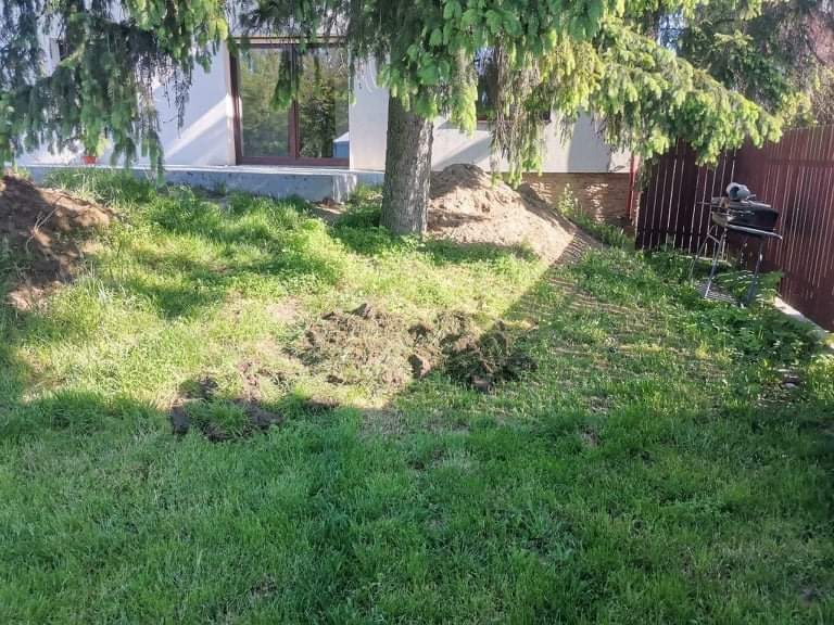 Teren przed domem z usuniętą darnią, pryzma ziemi przy pniu drzewa iglastego, w tle widoczny fragment budynku i ogrodzenie.
