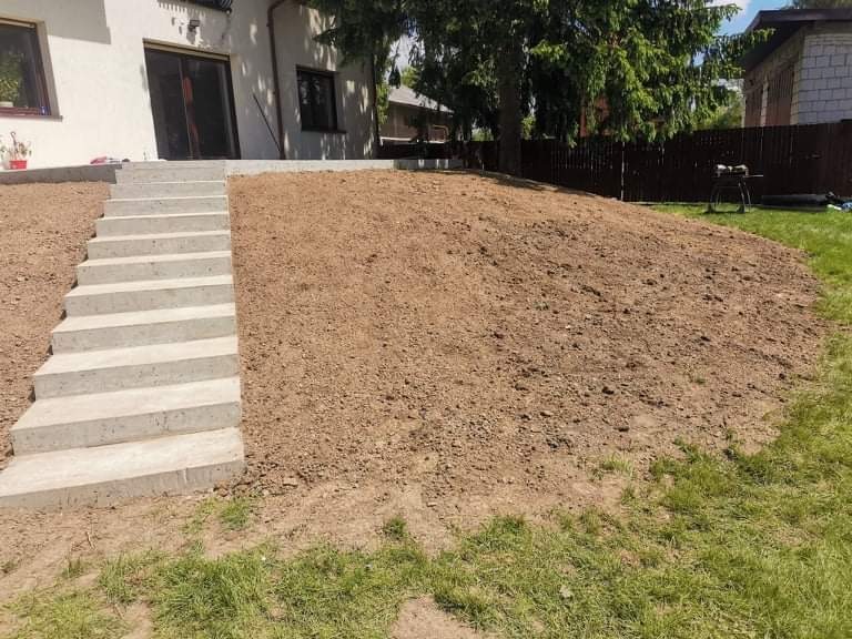 Przygotowany teren pod trawnik z betonowymi schodami prowadzącymi do domu, widoczna świeżo przekopana ziemia i fragment trawy.