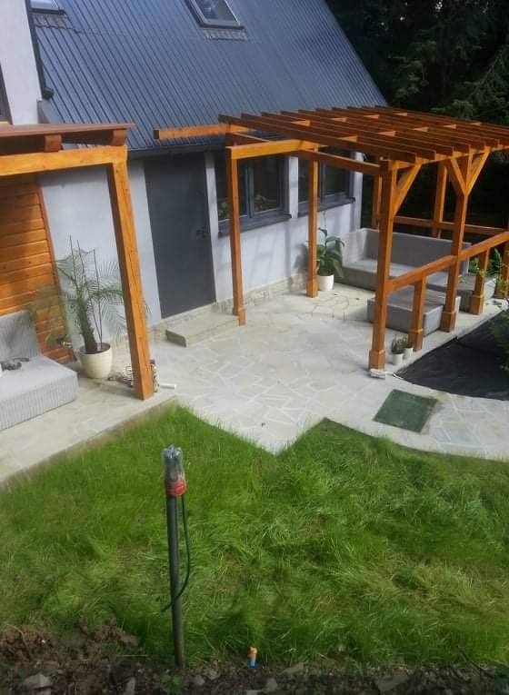 Wykończony taras z jasnej, nieregularnej kostki, z drewnianą pergolą i meblami ogrodowymi, przylegający do budynku z ciemnym dachem i fragmentem trawnika z widocznym słupkiem oświetleniowym.