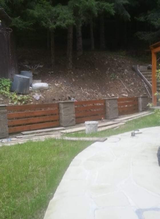 Nowo wybudowany murek oporowy z drewnianymi przęsłami i kamiennymi słupkami na tle zalesionego zbocza, widok z poziomu kamiennej ścieżki.