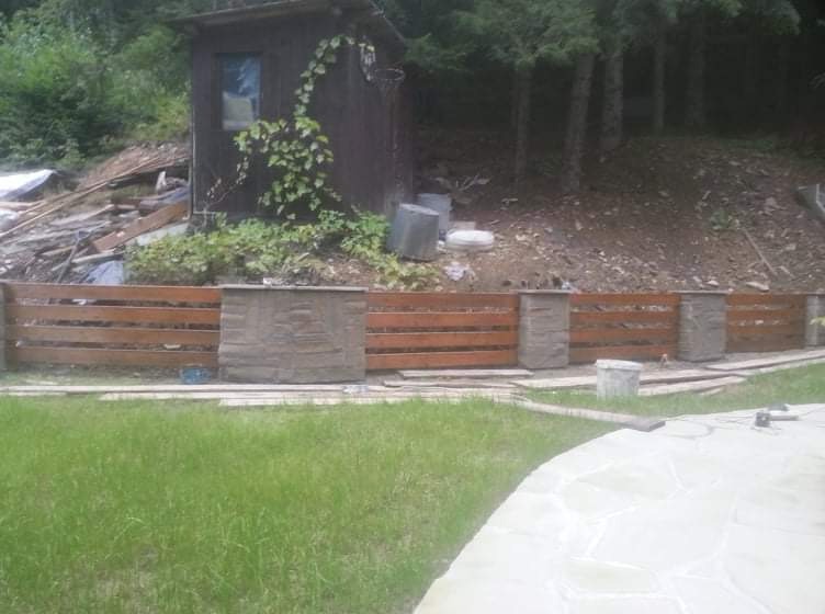 Nowo postawione drewniane ogrodzenie z betonowymi słupkami na nierównym terenie, z widocznym bałaganem na zboczu za nim i zielonym trawnikiem z przodu.