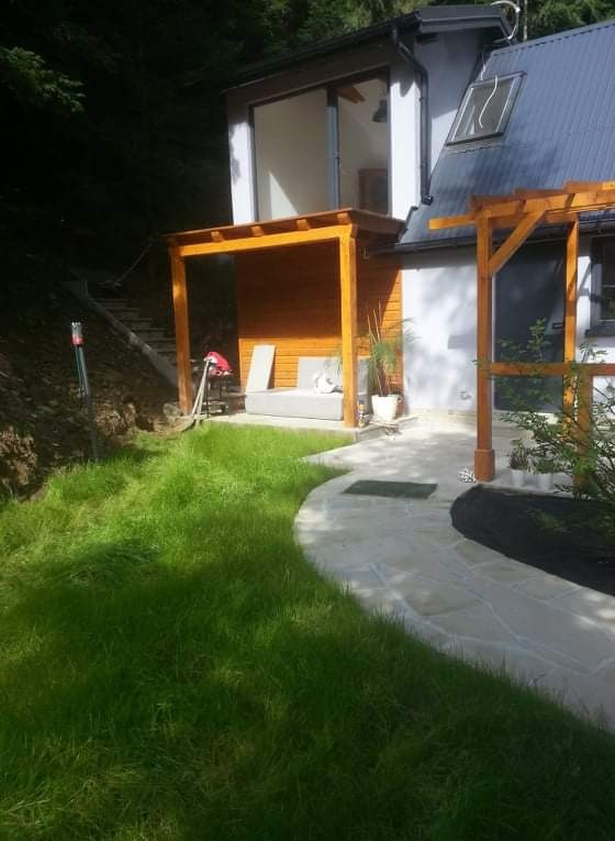 Widok na dom z ogrodem, z drewnianą pergolą i tarasem z kostki granitowej, trawa częściowo skoszona, na tarasie sofa z poduszkami, na trawniku leży szpadel.