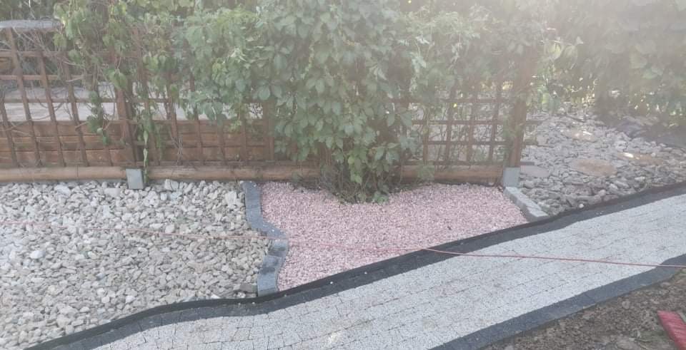 Niedokończony chodnik z szarej kostki brukowej z czarną obwódką, obok rabata z różowego żwiru i kamieni, w tle drewniany płot z pnączami.
