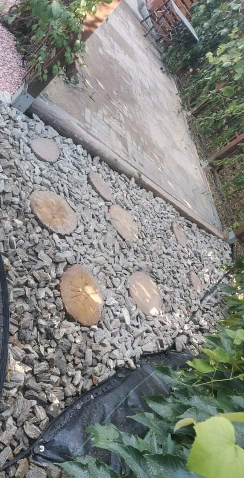 Ścieżka w ogrodzie wykonana z drewnianych krążków i szarego grysu, obok drewniana palisada i zielona roślinność.