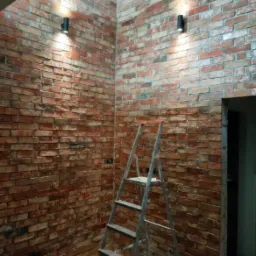 Kąt wewnętrzny pomieszczenia z wyeksponowaną ścianą z cegły klinkierowej, oświetloną dwoma czarnymi kinkietami, z rozstawioną aluminiową drabiną malarską.