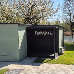 GM Box Sandra Grzegorzek Michał Machaczek s.c. - Nowoczesny budynek w szałwiowej zieleni z numerem 5 i czarną wnęką z neonem 'nova', otoczony zielenią, kwitnącym drzewem i widokiem na jezioro.