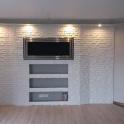 Ściana z dekoracyjnej białej cegły z wnękami i zamontowanym telewizorem, oświetlona punktowymi lampami sufitowymi, widok na podłogę z paneli i częściowo widoczne drzwi.