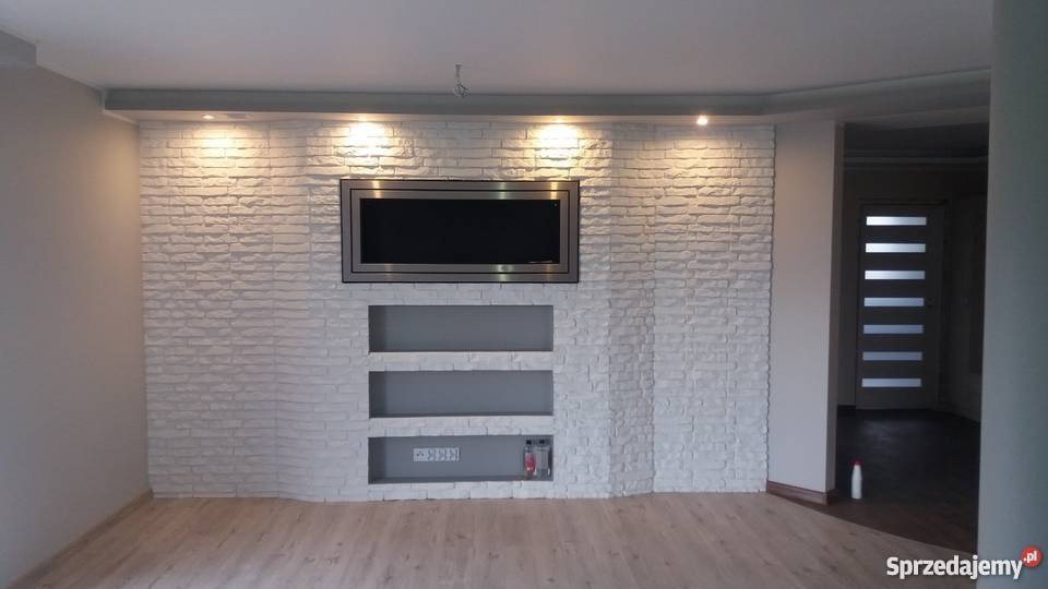 Ściana z dekoracyjnej białej cegły z wnękami i zamontowanym telewizorem, oświetlona punktowymi lampami sufitowymi, widok na podłogę z paneli i częściowo widoczne drzwi.