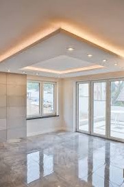 Jasne wnętrze z marmurową posadzką, dużymi oknami i podwieszanym sufitem z oświetleniem LED w kształcie trójkąta i kwadratu, odbijającym się w polerowanej podłodze.