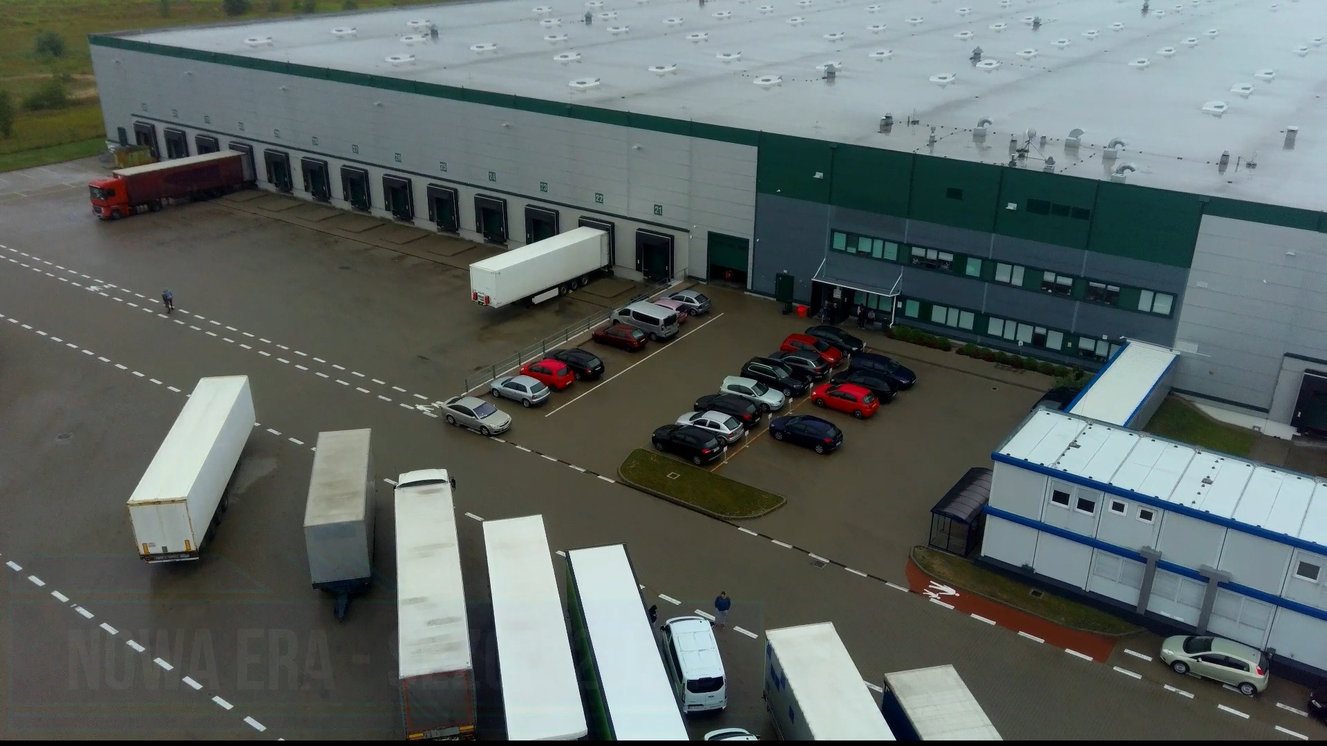 Widok z góry na parking i rampy rozładunkowe dużego magazynu logistycznego z zaparkowanymi samochodami osobowymi i ciężarówkami z naczepami.