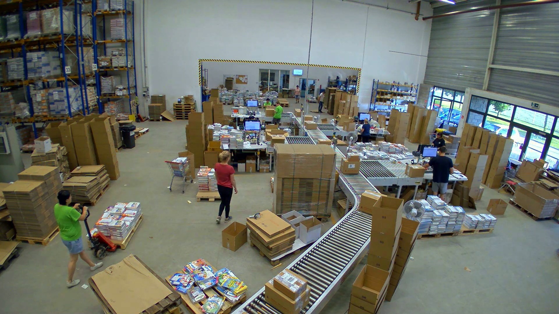 Szeroki kadr z góry na dużą halę magazynową z pracownikami przy stołach, konfekcjonującymi i pakującymi towary na paletach oraz na taśmach produkcyjnych, przygotowując je do wysyłki.