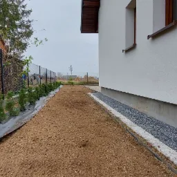 Przygotowany teren pod trawnik i nasadzenia tujek przy ogrodzeniu z siatki i obok ściany domu z oknami. Widoczny fragment chodnika z kostki brukowej.