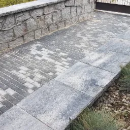 Ułożona z szarej kostki brukowej ścieżka prowadząca do metalowej bramy, ograniczona z jednej strony kamiennym murkiem z wystającymi elementami przypominającymi korzenie, a z drugiej strony niskim...