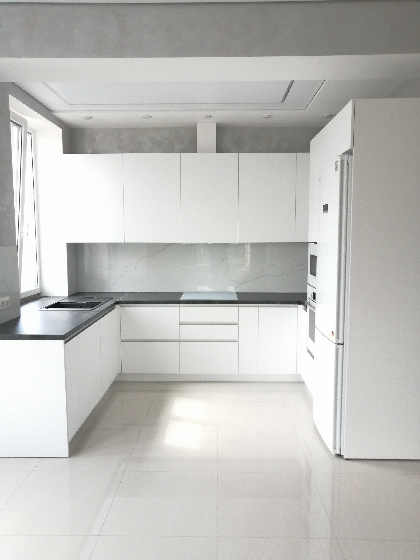 Minimalistyczna, nowoczesna kuchnia z białymi szafkami i ciemnym blatem, marmurowy backsplash, jasna podłoga, oświetlenie punktowe.