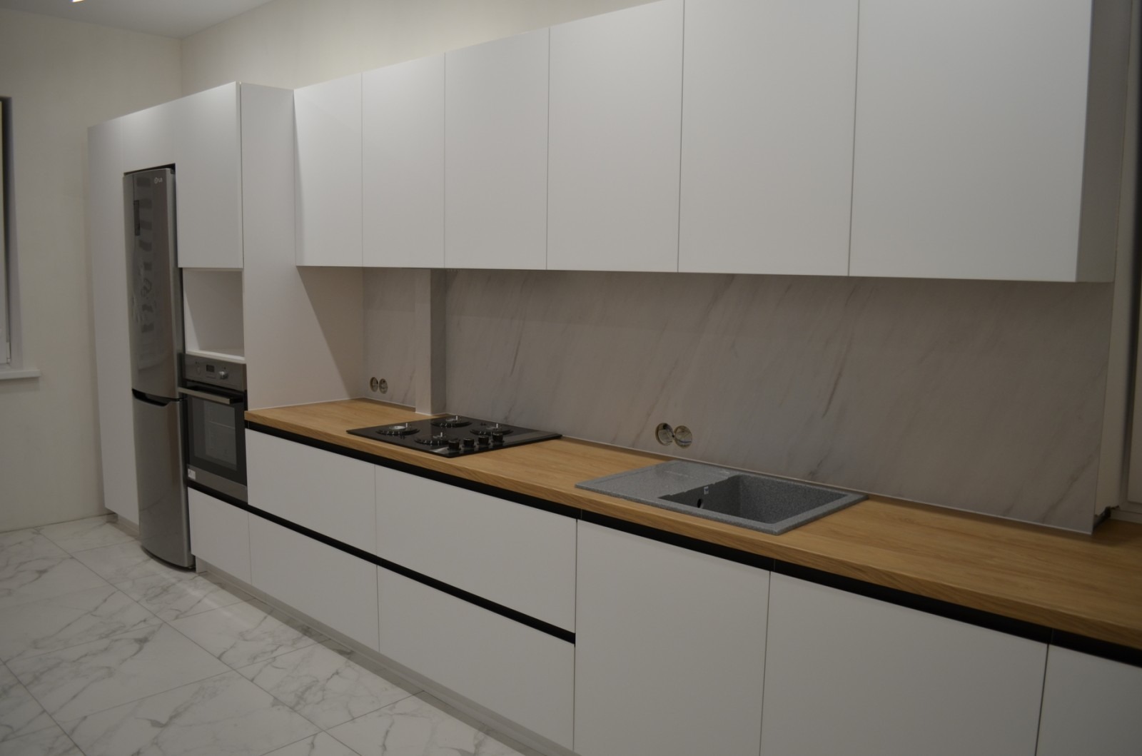 Nowoczesna kuchnia z białymi szafkami, drewnianym blatem i marmurowym backsplashiem, z widoczną płytą grzewczą i szarym zlewem.