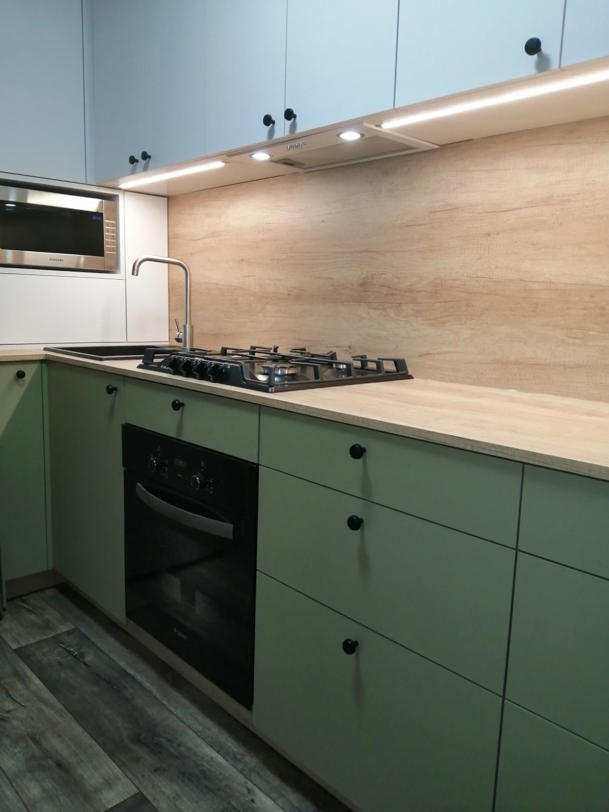 Nowoczesna kuchnia z zielonymi szafkami, drewnianym panelem nad blatem, czarną płytą gazową i piekarnikiem, oraz srebrnym kranem i mikrofalówką.