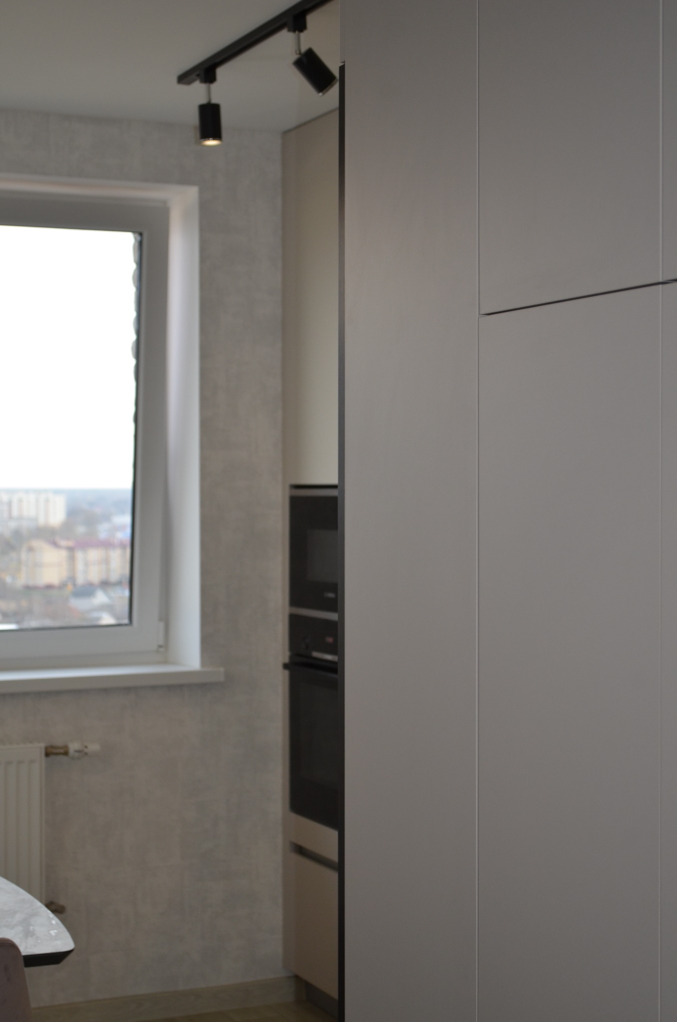 Minimalistyczna kuchnia z szarymi frontami szafek, czarnym piekarnikiem i mikrofalówką oraz punktowym oświetleniem na szynie, widok z okna na miasto w tle.