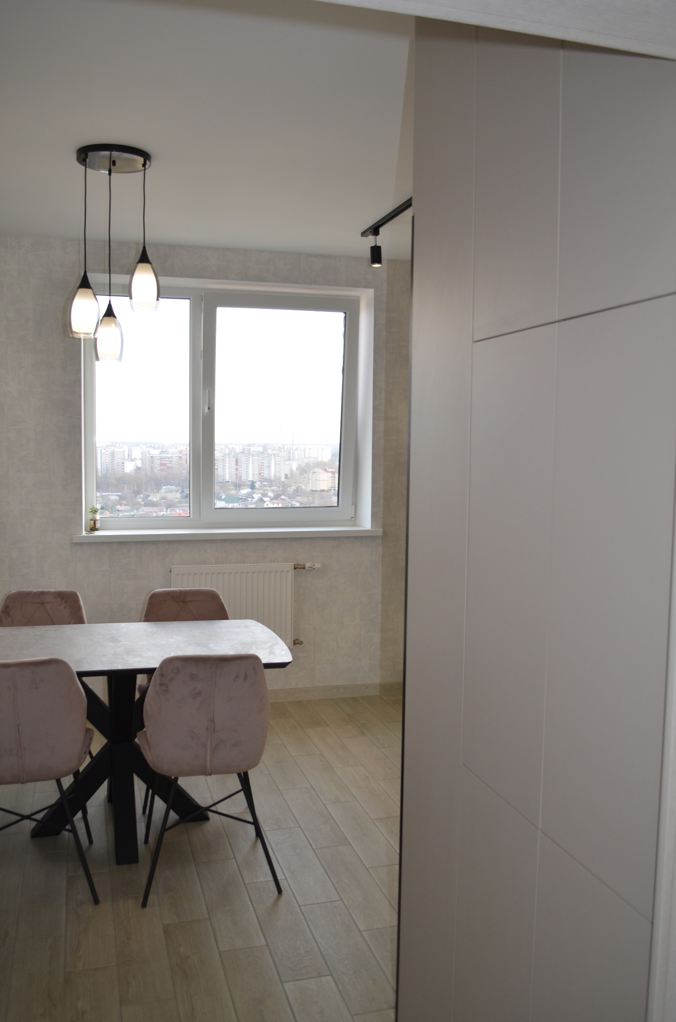 Jasna jadalnia z drewnianą podłogą, stołem z czarną podstawą i różowymi krzesłami, wiszącą lampą z trzema kloszami oraz widokiem na miasto za oknem, częściowo przysłonięta białym panelem.