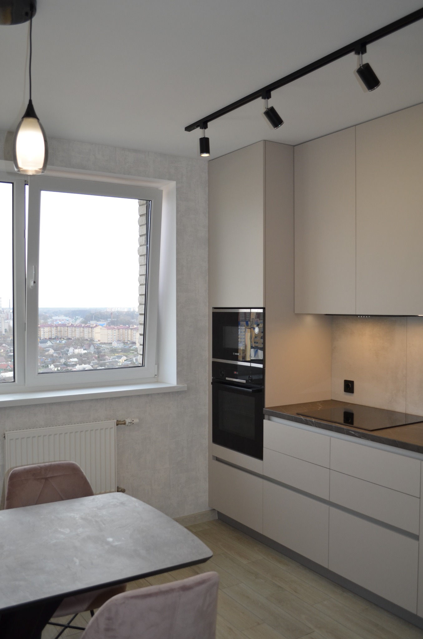 Jasna kuchnia z jadalnią w Warszawie: minimalistyczne, beżowe szafki kuchenne, czarny piekarnik i płyta indukcyjna, stół z szarym blatem i różowe krzesła, oświetlenie punktowe na suficie, widok...