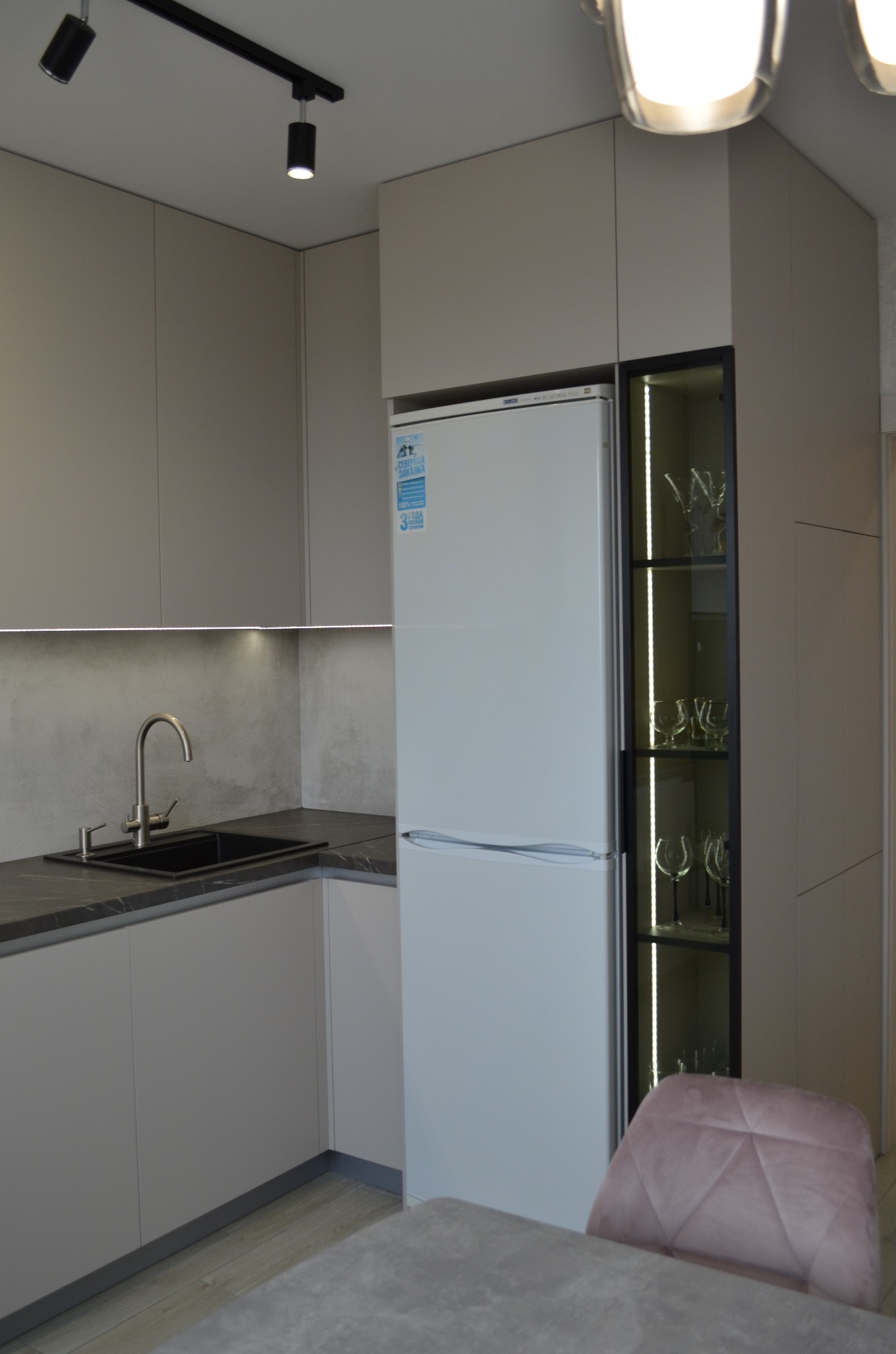 Nowoczesna kuchnia w odcieniach szarości z czarnym zlewem i marmurowym blatem, oświetleniem podszafkowym LED i wysoką zabudową z lodówką oraz przeszkloną witryną z kieliszkami, krzesło z różowego...