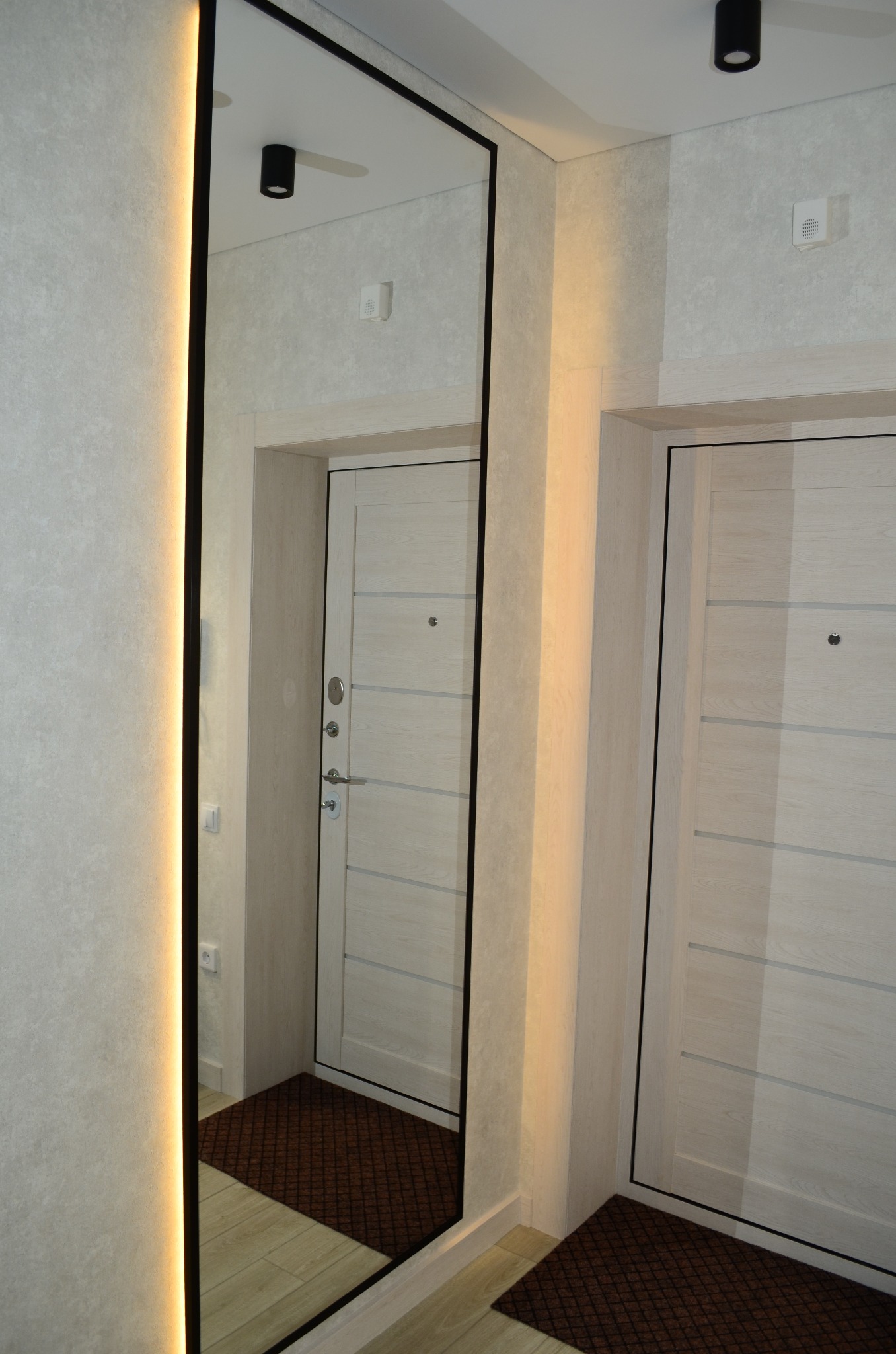 Nowoczesny przedpokój z dwoma białymi drzwiami, dużym lustrem z podświetleniem LED i brązową wycieraczką