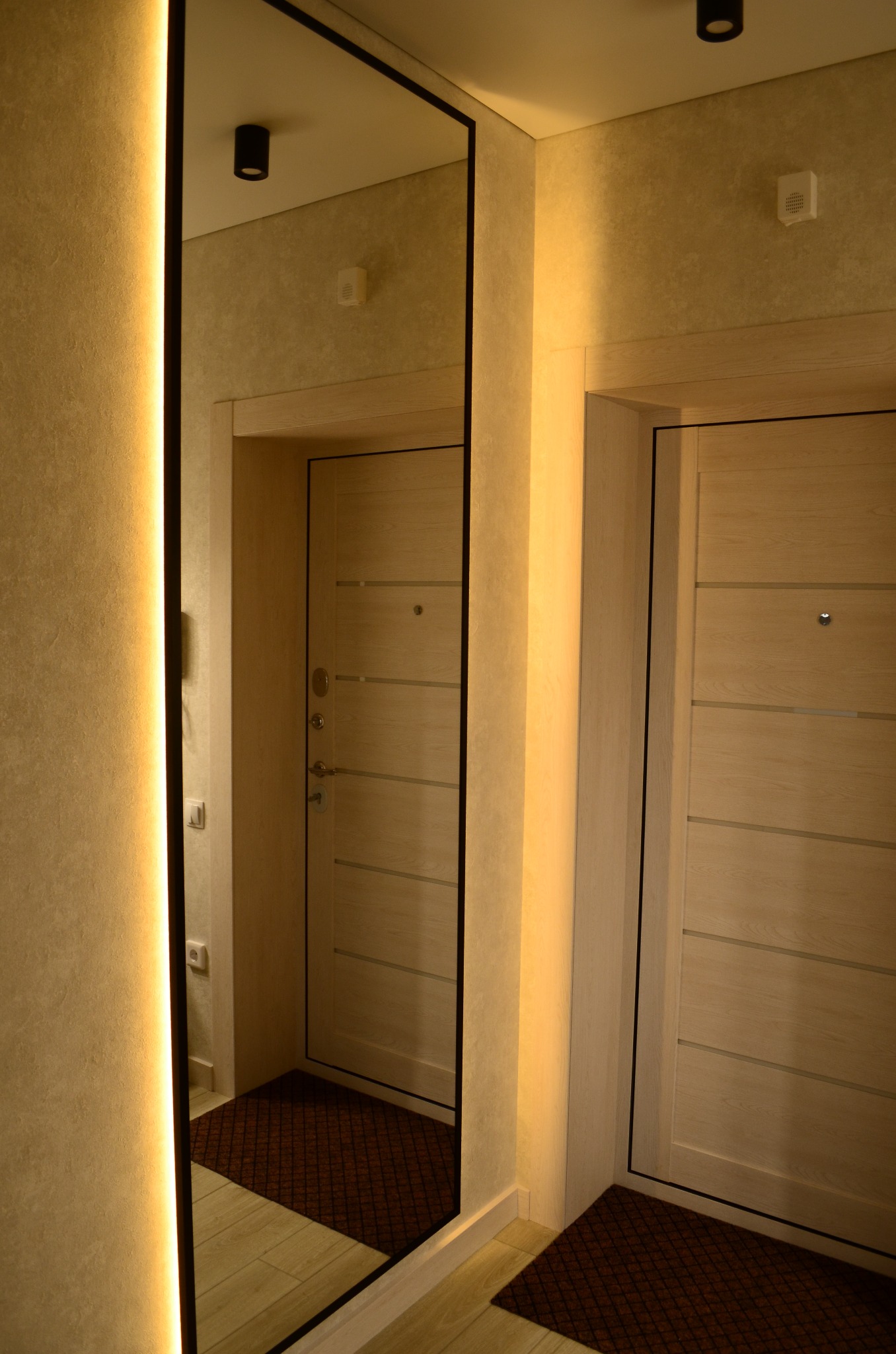 Nowoczesny przedpokój z lustrem w czarnej ramie, podświetleniem LED i dwiema jasnymi drewnianymi drzwiami wejściowymi.