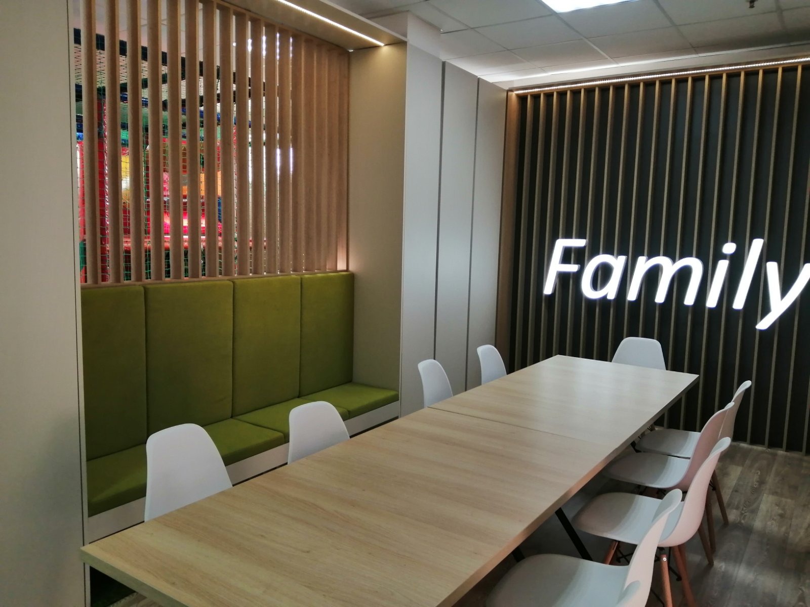Nowoczesna sala konferencyjna z długim stołem, białymi krzesłami i zieloną wnęką z siedziskami, podświetlonym napisem 'Family' na ścianie z drewnianych listew.