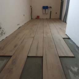 WykończMnie - Układanie podłogi z paneli imitujących drewno w pomieszczeniu w trakcie remontu; widoczne fragmenty surowego podłoża i ślady po pracach na ścianie.