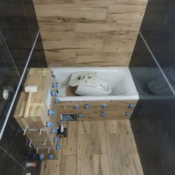 WykończMnie - Układanie płytek imitujących drewno w łazience, widoczne krzyżyki glazurnicze, wanna obudowana płytkami, fragment instalacji elektrycznej.