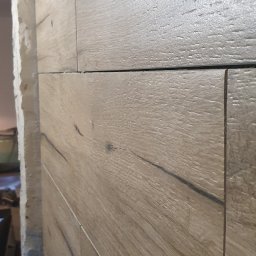 WykończMnie - Ściana wykończona panelami imitującymi drewno, widoczny fragment ściany z otworem i szczelinami między panelami, niedokończone wykończenie przy framudze drzwi.