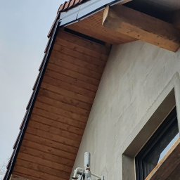 WykończMnie - Fragment elewacji budynku z widocznym oknem, drewnianą podbitką dachu, fragmentem rusztowania i ociepleniem w trakcie prac wykończeniowych.