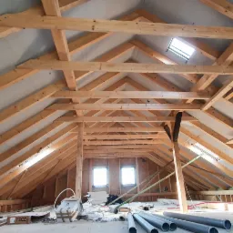 Konstrukcja drewniana dachu poddasza z oknami dachowymi i kominem murowanym z bloczków betonowych, widoczne rury PCV i materiały budowlane na podłodze.