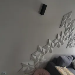 Ściana w salonie z dekoracyjnymi trójwymiarowymi panelami w kształcie rombów, rozmieszczonymi asymetrycznie, podświetlona dwoma czarnymi reflektorami, z widocznym fragmentem szarej sofy i oknem...