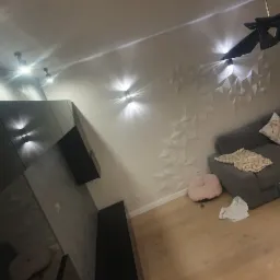 Nowoczesny salon z szarą sofą narożną, szklanym stolikiem kawowym, dekoracyjną ścianą z trójwymiarowych paneli i oświetleniem punktowym.