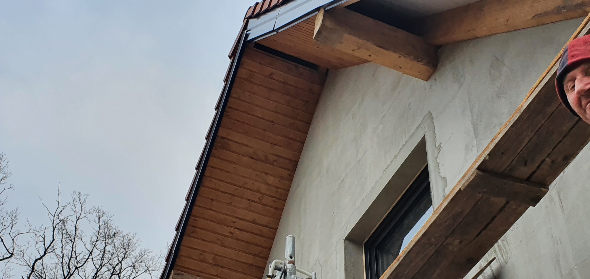 Fragment elewacji budynku z widocznym oknem, drewnianą podbitką dachu, fragmentem rusztowania i ociepleniem w trakcie prac wykończeniowych.