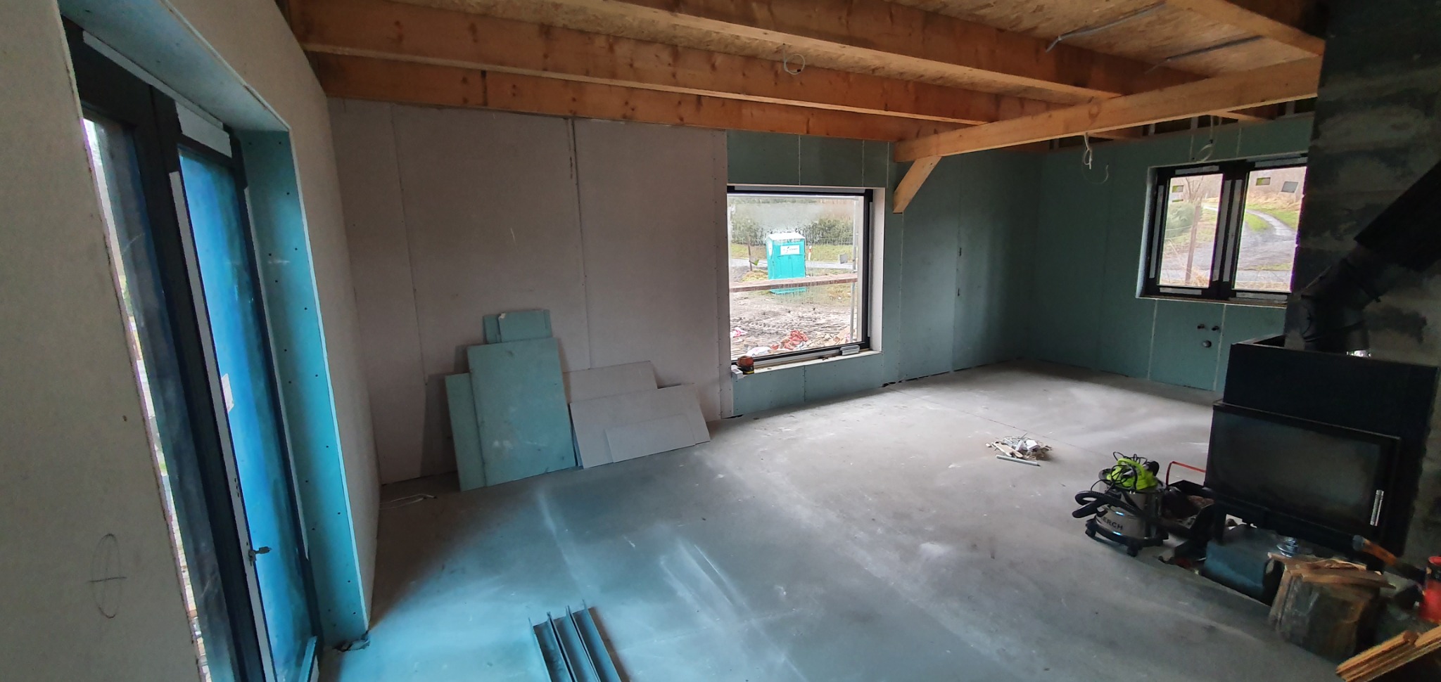 Wnętrze surowego domu w trakcie wykańczania, ściany pokryte zielonkawymi płytami, widoczne drewniane belki stropowe, okna z widokiem na zewnątrz, na podłodze resztki materiałów budowlanych...