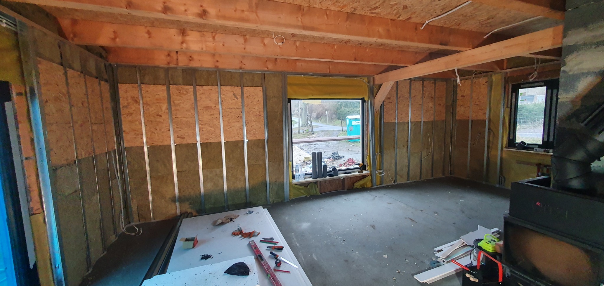 Wnętrze w trakcie prac wykończeniowych: ściany z izolacją, metalowy stelaż pod płyty, surowy drewniany strop, okna z widokiem na zewnątrz, narzędzia na stole, betonowa podłoga.