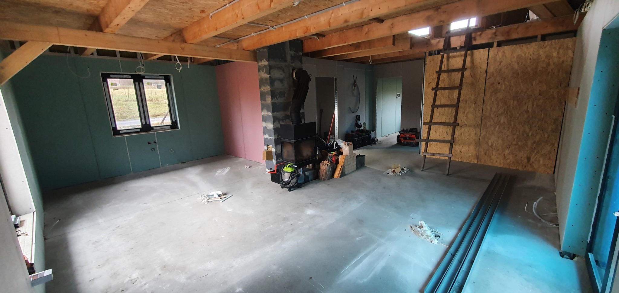 Wnętrze w trakcie wykańczania: ściany pokryte zielonymi płytami, widoczne drewniane belki stropowe, kominek, drabina oparta o ścianę z płyty OSB, podłoga betonowa, okno z widokiem na zewnątrz.