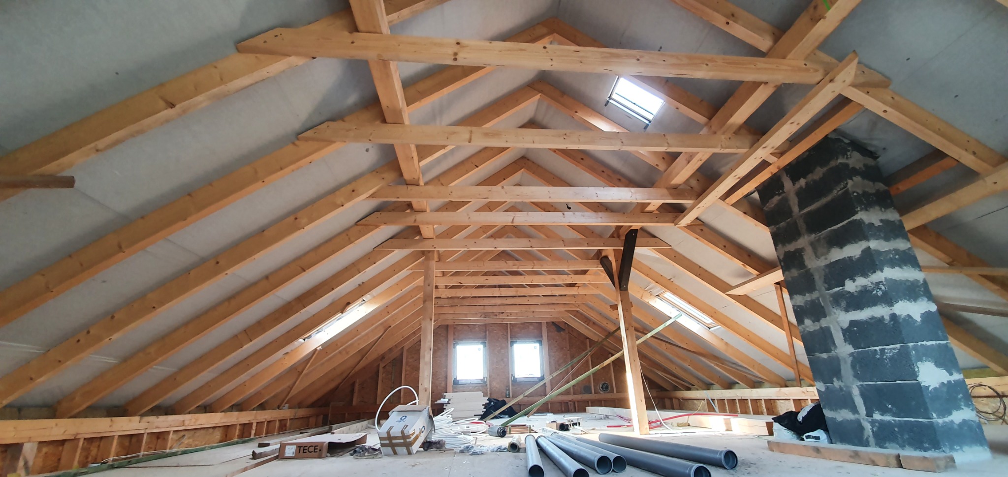 Konstrukcja drewniana dachu poddasza z oknami dachowymi i kominem murowanym z bloczków betonowych, widoczne rury PCV i materiały budowlane na podłodze.