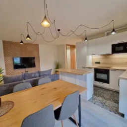 Jasne wnętrze: salon z szarą sofą i TV na tle drewnianej ściany, otwarta kuchnia z wyspą, drewniany stół z szarymi krzesłami i dekoracyjne lampy wiszące.