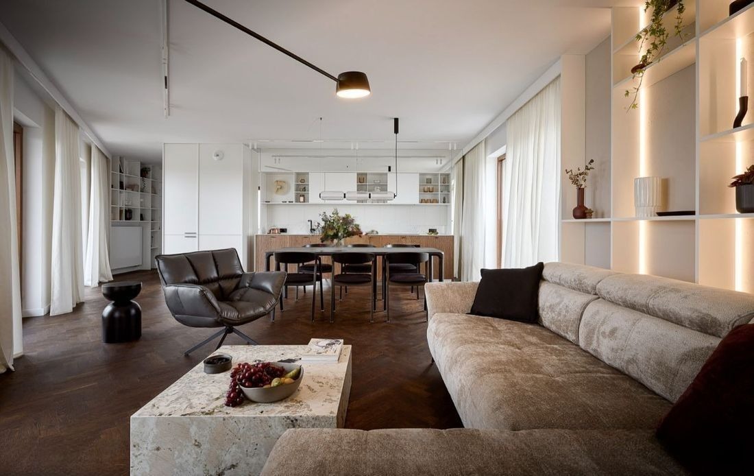 Jasne, przestronne wnętrze mieszkania z parkietem w jodełkę, beżową sofą i marmurowym stolikiem kawowym. W tle widoczna kuchnia i jadalnia w stylu minimalistycznym.