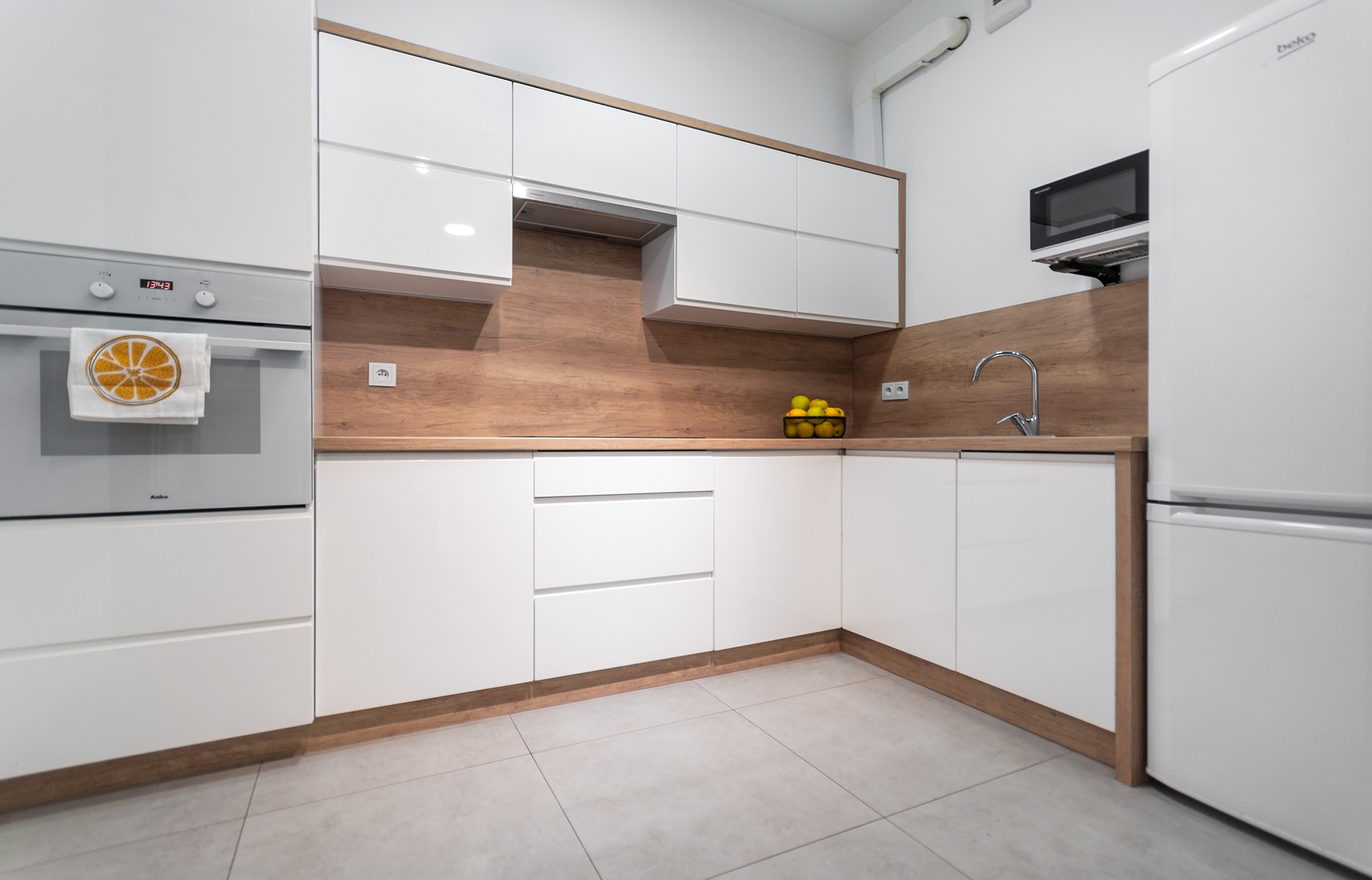 Nowoczesna kuchnia z białymi szafkami i drewnianym blatem. Na blacie koszyk z cytrynami. Na piekarniku ręcznik z motywem pomarańczy. Minimalistyczny design.