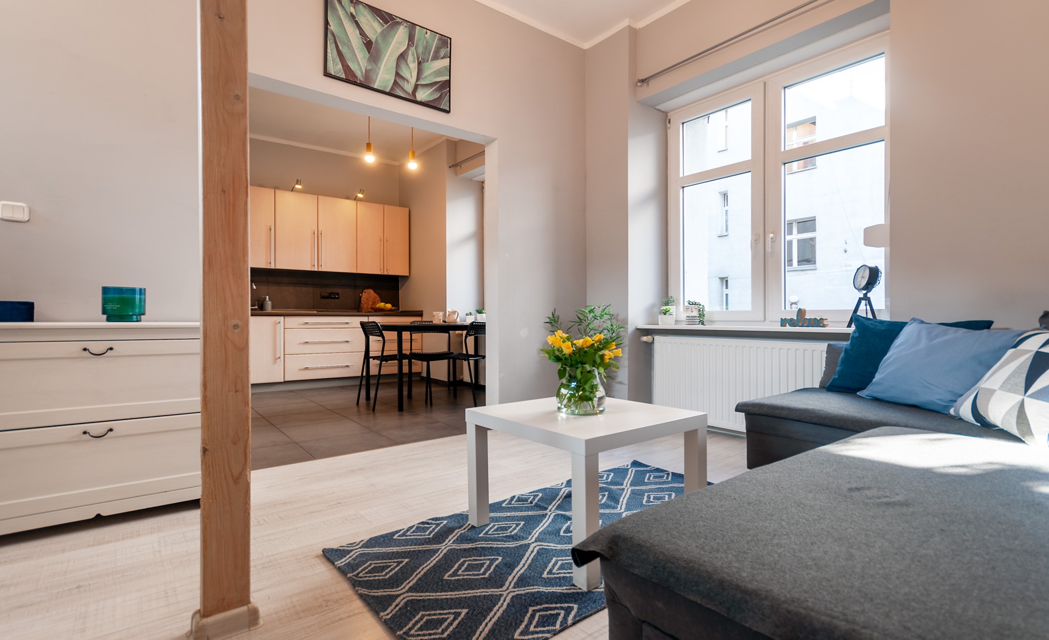 Jasne wnętrze mieszkania z otwartą kuchnią, kanapą i dywanem w geometryczne wzory. Na stole wazon z żółtymi kwiatami. Widok na budynek z okna.