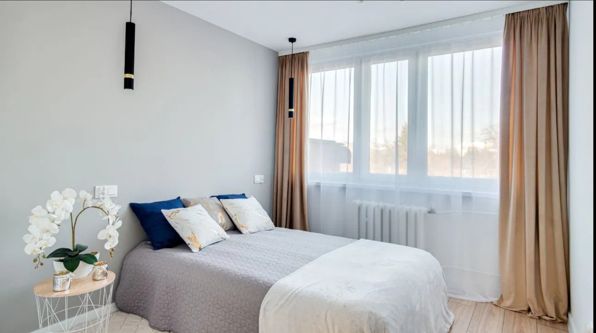 Jasna sypialnia z łóżkiem, błękitnymi poduszkami i kwiatami na stoliku nocnym. Okno z beżowymi zasłonami wpuszcza naturalne światło.