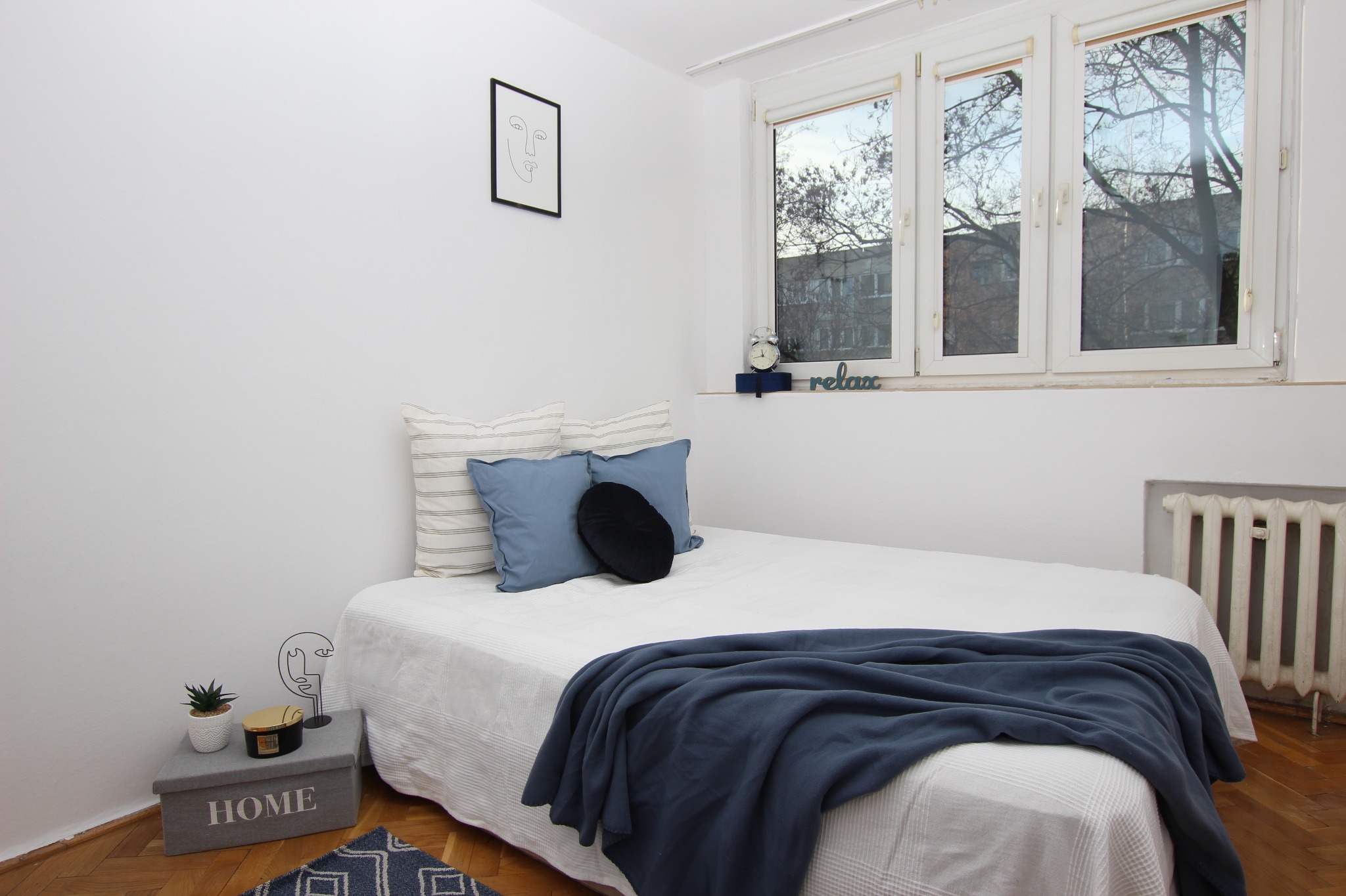 Jasne wnętrze sypialni z łóżkiem, niebieskimi poduszkami i pledem. Na ścianie minimalistyczny obrazek, a na szafce nocnej dekoracje i roślina w doniczce. Parkiet na podłodze.