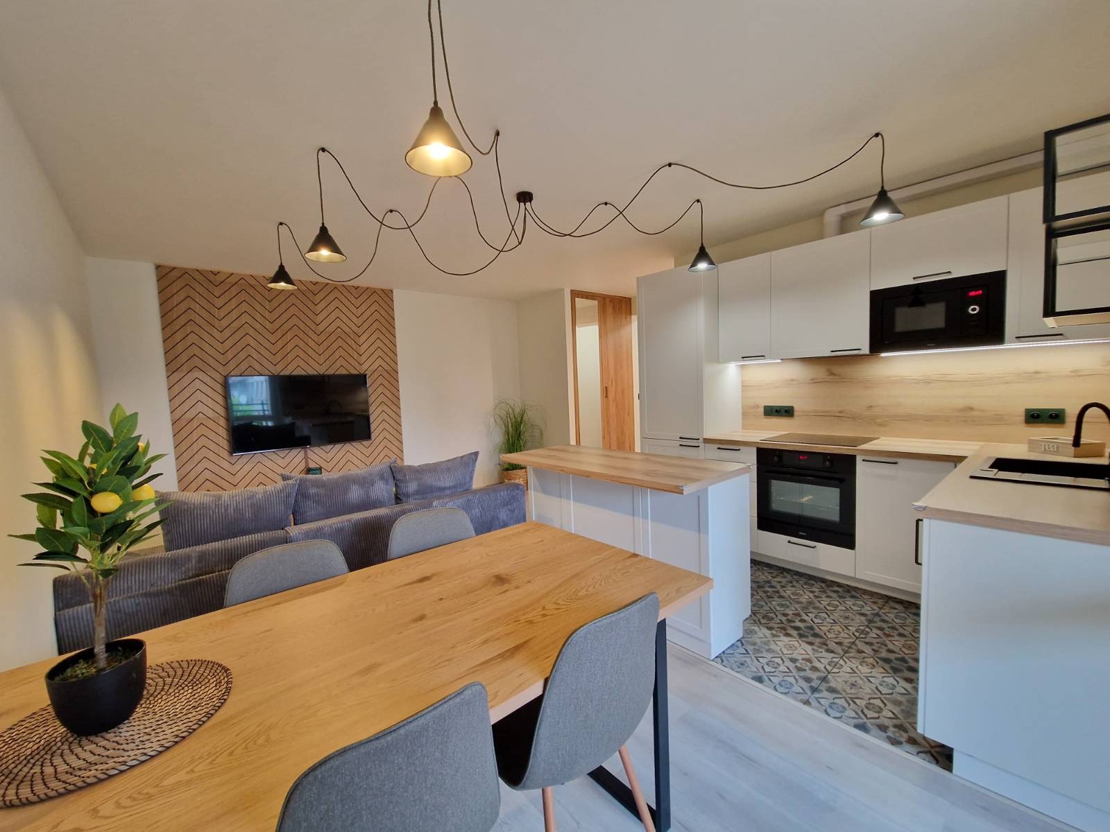 Jasne wnętrze: salon z szarą sofą i TV na tle drewnianej ściany, otwarta kuchnia z wyspą, drewniany stół z szarymi krzesłami i dekoracyjne lampy wiszące.