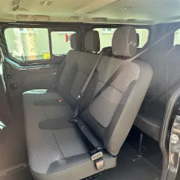 Wnętrze czarnego busa z trzema rzędami szarych siedzeń, widok z boku na pasy bezpieczeństwa i okna z widokiem na budynek.