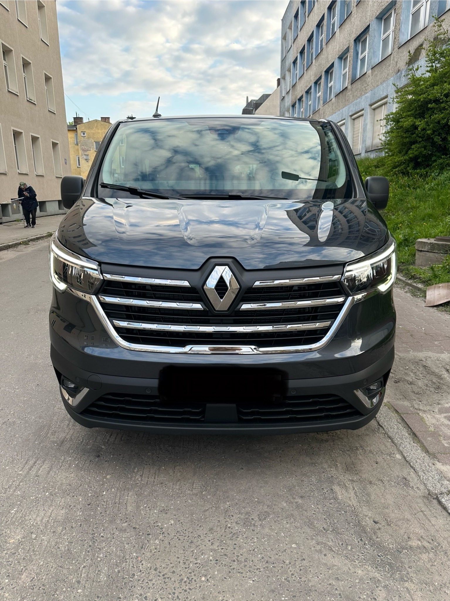 Przód szarego busa Renault z chromowanymi elementami na tle miejskiego krajobrazu z budynkami i chodnikiem.