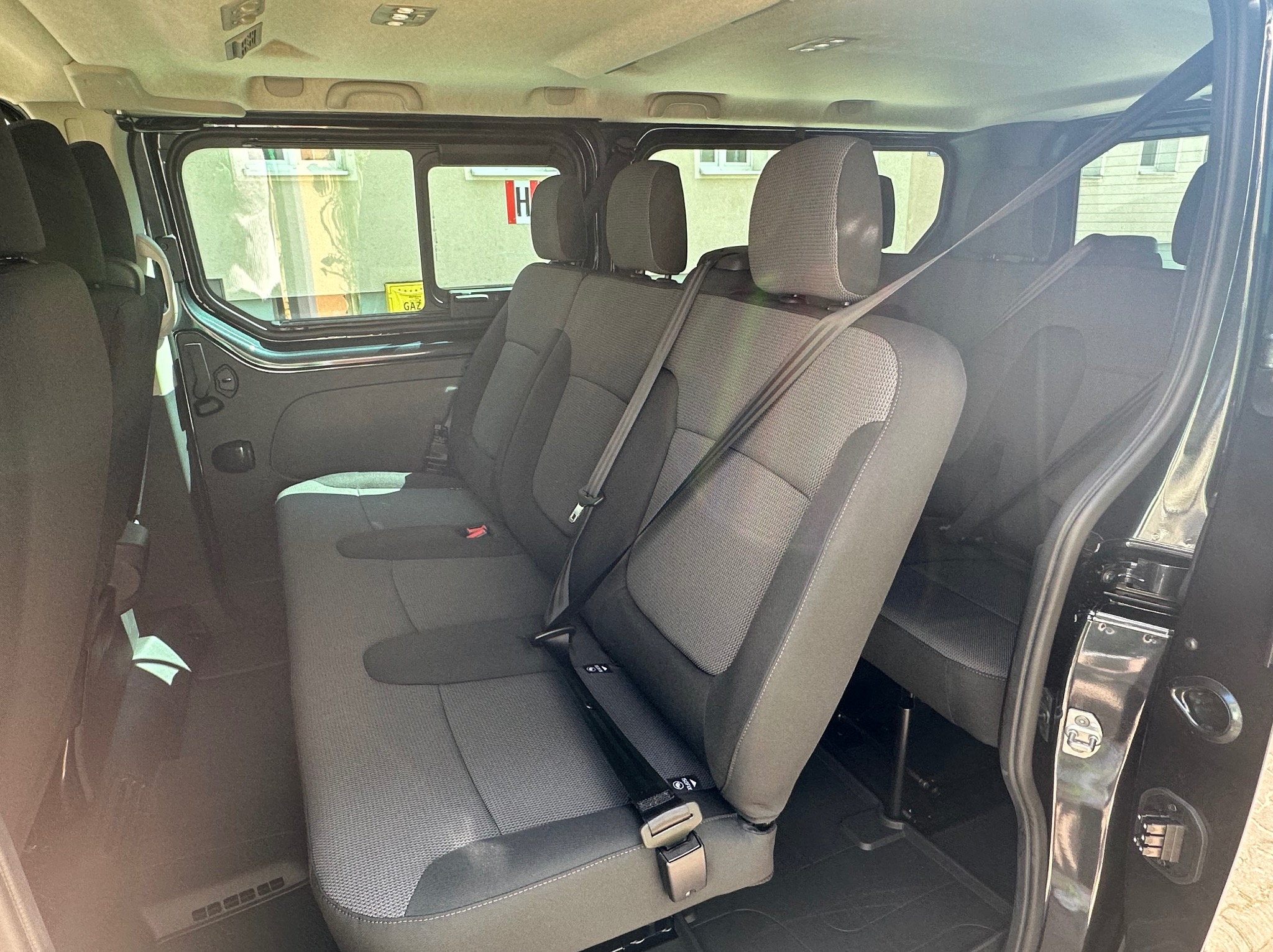 Wnętrze czarnego busa z trzema rzędami szarych siedzeń, widok z boku na pasy bezpieczeństwa i okna z widokiem na budynek.