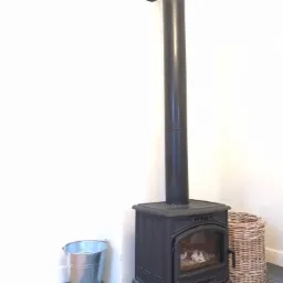 Czarny piec wolnostojący z rurą odprowadzającą dym, obok ocynkowane wiadro i wiklinowy kosz na tle białej ściany i podłogi w szarą szachownicę.